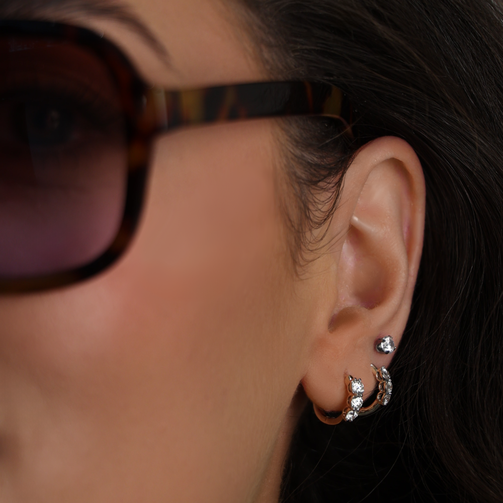 Close-up of an ear with 2 pairs of diamond hoop earrings, one stud earring and sunglasses on a blurred background